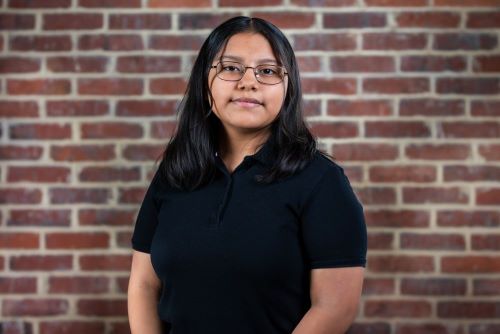 Melany Mendez Casas, a young Latina woman, stands in front of a brick wall. She wears glasses and a black polo shirt.