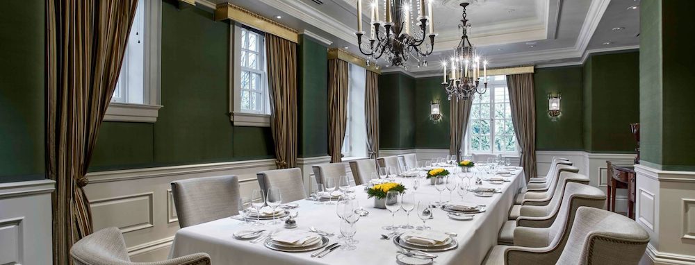 The image shows an elegantly decorated dining room with a long table set for a formal dinner, featuring a white tablecloth, neatly arranged plates, glasses, and cutlery, with chandeliers hanging from the ceiling and large windows draped with curtains.