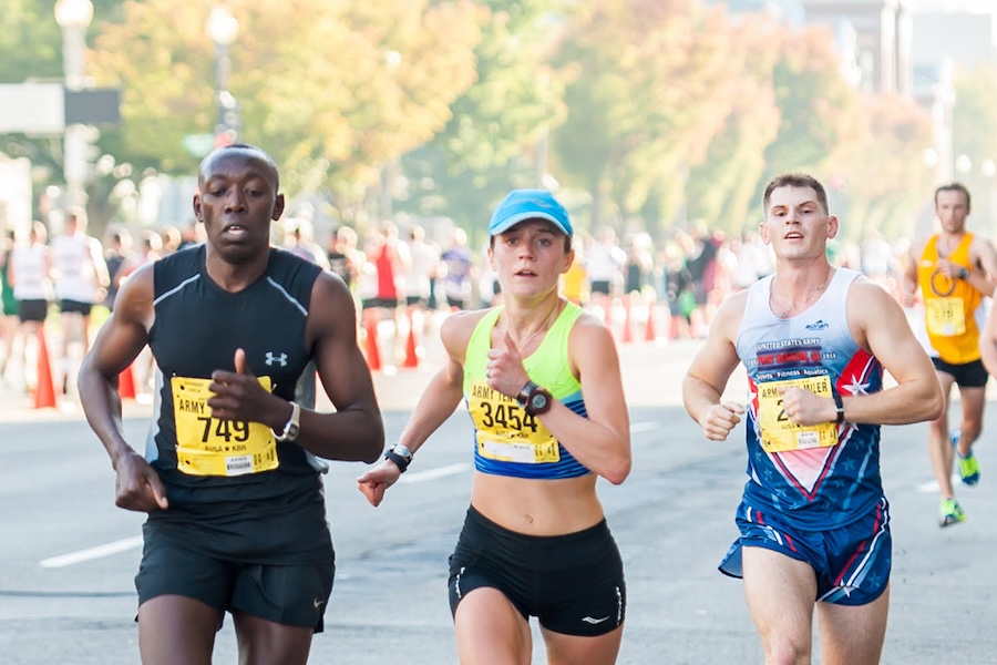 A group of competitive runners in a race on a city street. The runners are wearing various race bibs and athletic gear, with spectators and more participants visible in the background.