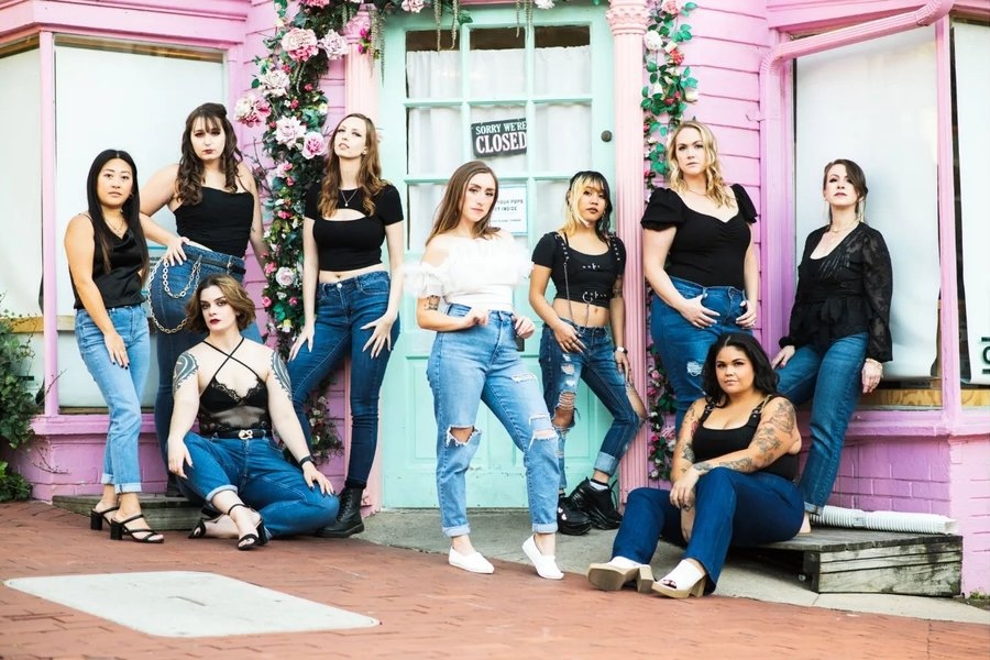 A group of women in coordinated outfits pose confidently in front of a pink building with a 'Sorry We're Closed' sign on the door.