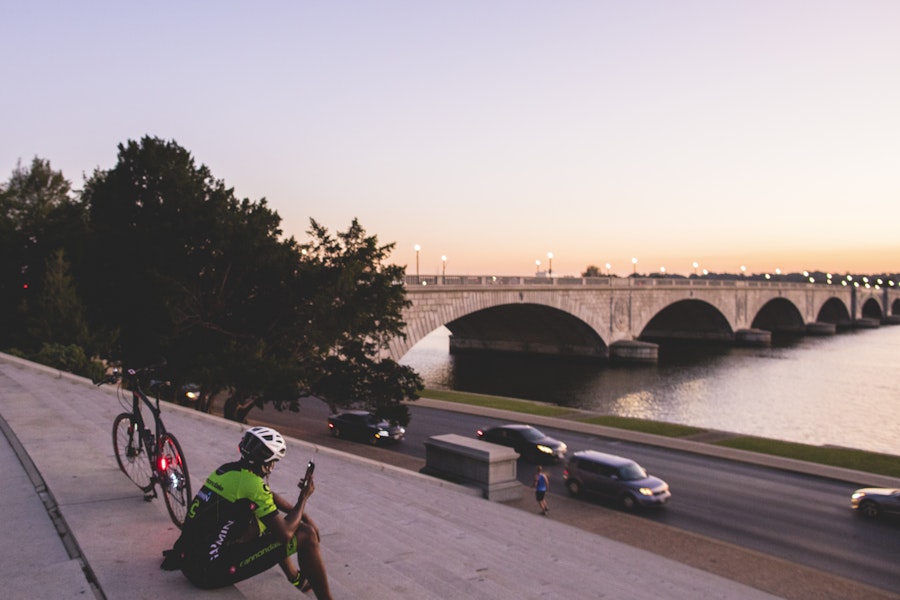 A cyclist sits on steps near the water, with their bike beside them, overlooking the Arlington Memorial Bridge at sunset.