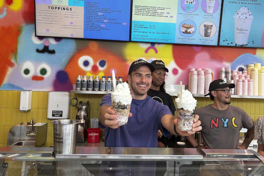 A smiling man behind an ice cream counter holds out two large milkshakes topped with whipped cream. He is flanked by two other employees, and a brightly colored, playful mural is visible behind them, along with a menu board overhead.