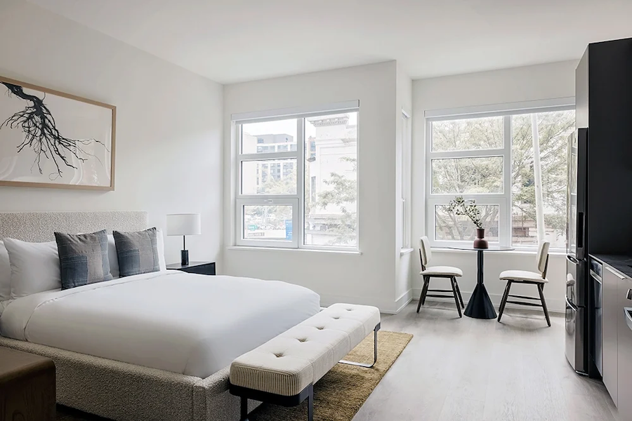 A bright, modern hotel room with a king-sized bed, a cushioned bench at the foot of the bed, and a small dining table with two chairs by the window. Large windows let in natural light, and the room is decorated in neutral tones with minimalist artwork on the wall.