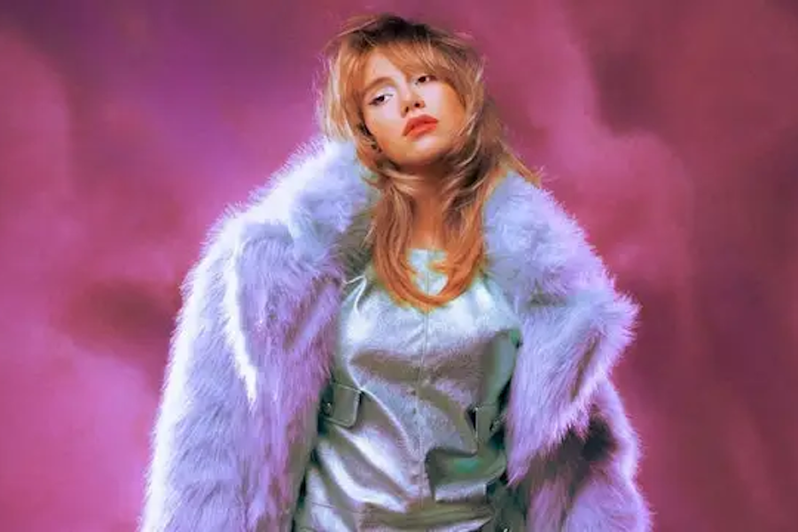 A woman with tousled blonde hair stands against a vibrant purple background, wearing a light blue outfit with a matching fluffy coat draped over her shoulders, giving a stylish and confident pose.