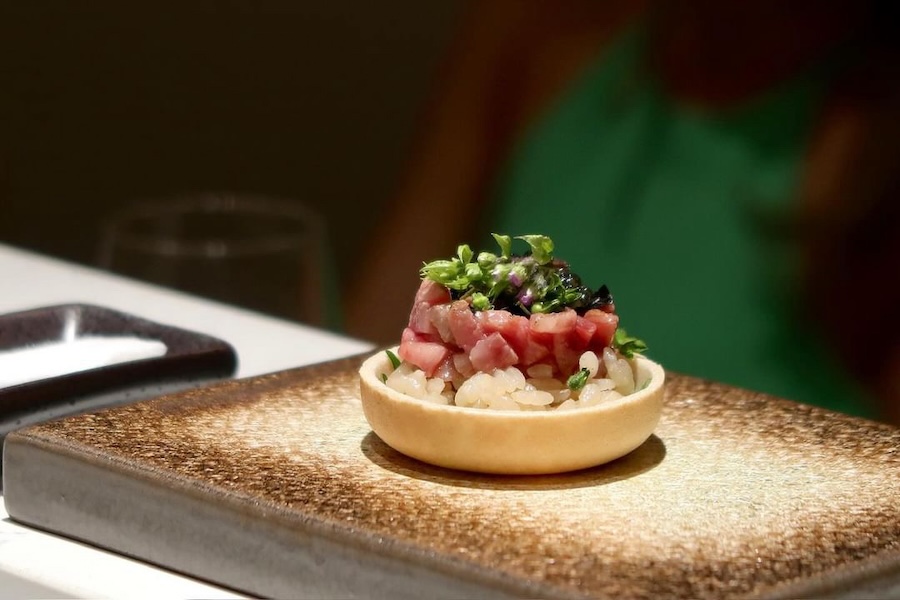 An intricately crafted tart topped with diced fish and herbs, served on a textured plate.