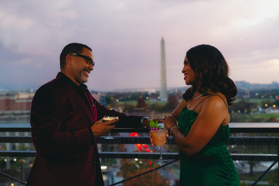 A couple enjoys cocktails on a rooftop terrace at sunset with the Washington Monument visible in the background.