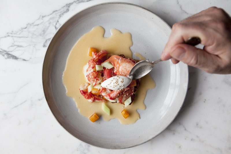 A saucy lobster dish on a white ceramic plate and marble table being broken into by a hand with a fork. 