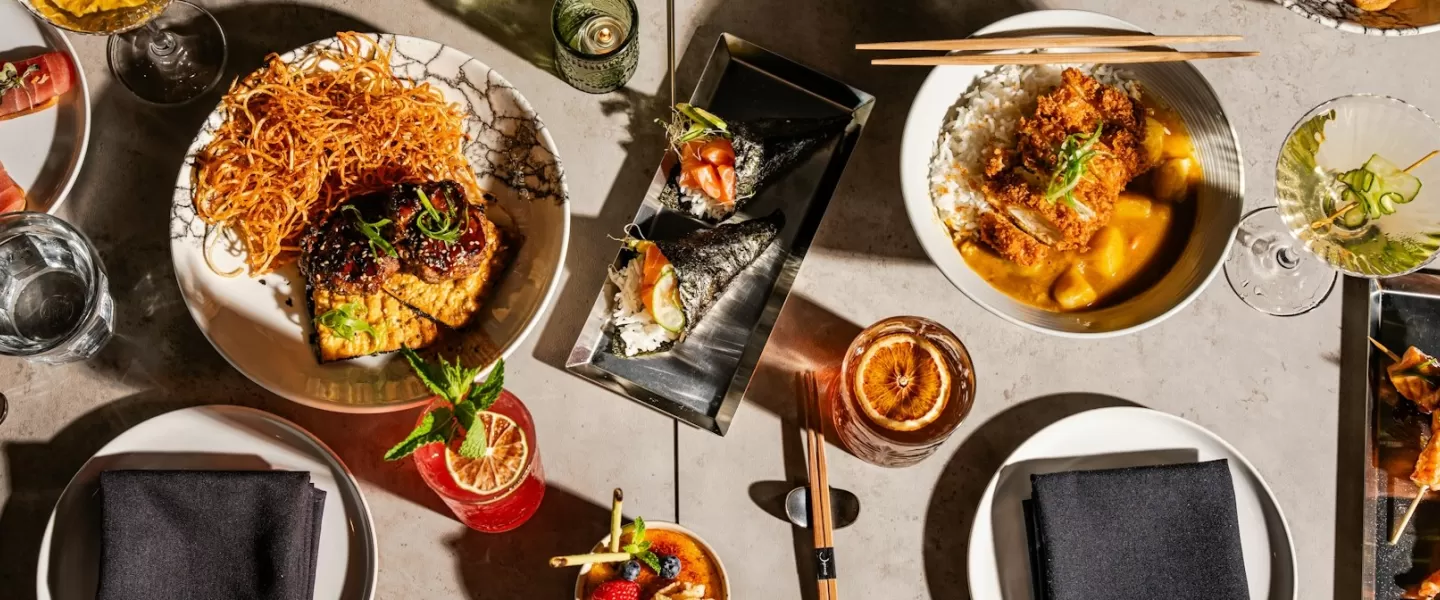 An overhead view of a diverse spread of Japanese dishes, featuring sushi rolls, rice bowls, and various appetizers, along with vibrant cocktails and meticulously plated dishes on a modern table setting.