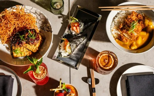 An overhead view of a diverse spread of Japanese dishes, featuring sushi rolls, rice bowls, and various appetizers, along with vibrant cocktails and meticulously plated dishes on a modern table setting.
