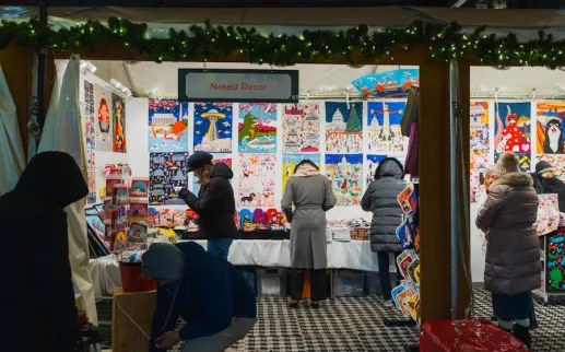 Shoppers browse colorful prints and gifts at the Naked Decor booth during a nighttime holiday market.
