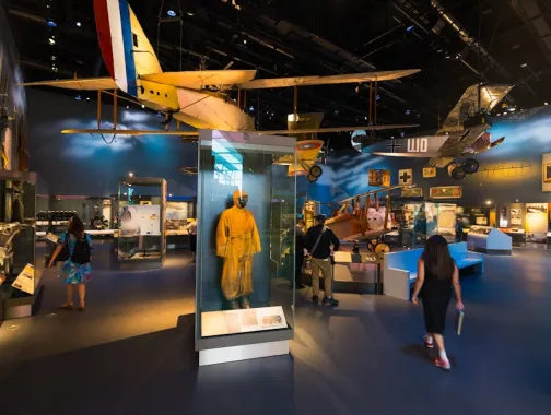 Visitors walk through an aviation exhibit with vintage aircraft and a yellow flight suit on display.