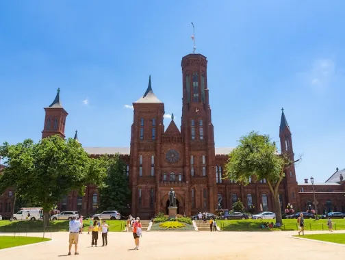 Visitors walk across the National Mall toward the red sandstone Smithsonian Castle on a sunny day.